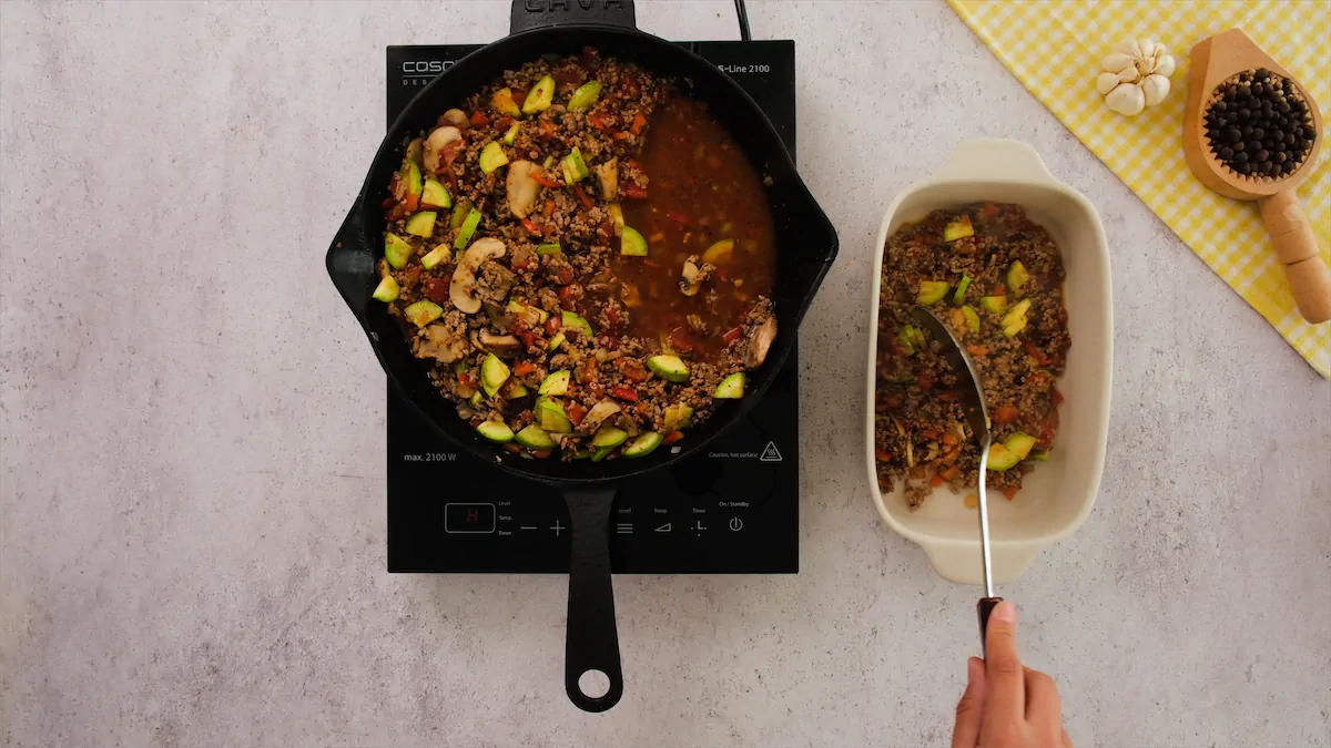 Ground beef with vegetables getting transferred from cast iron skillet to a baking dish with a spatula.
