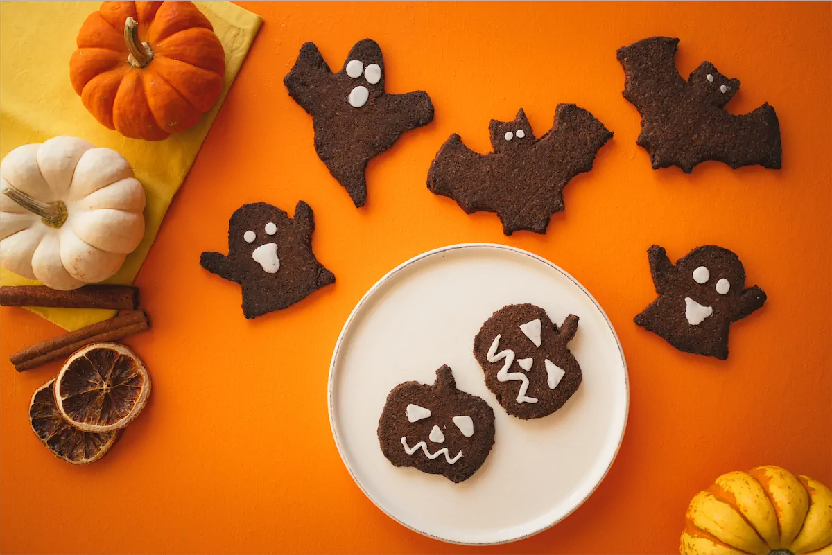 Bat and ghost-shaped keto Halloween cookies are arranged on an orange table alongside a plate of pumpkin shaped Halloween cookies.