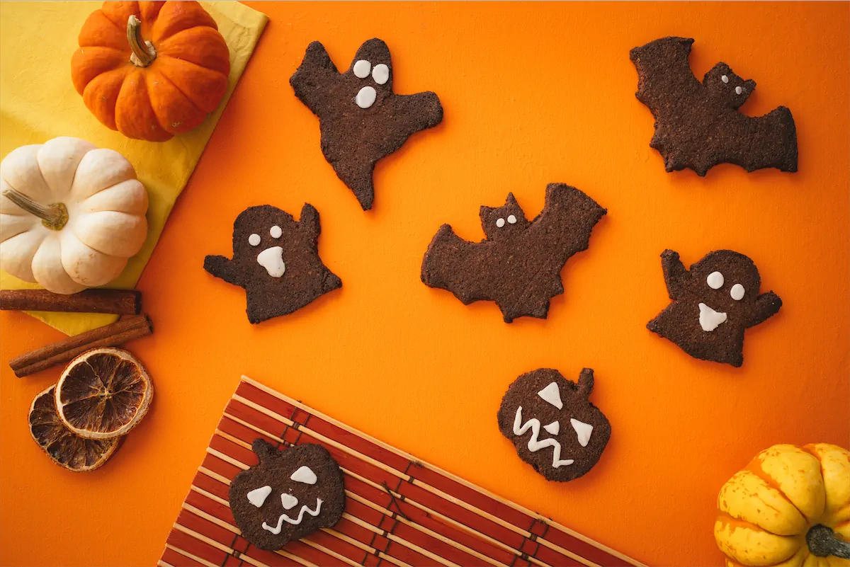 Bat, ghost, and pumpkin-shaped fun Halloween cookies are arranged on an orange table alongside red and yellow miniature pumpkins.