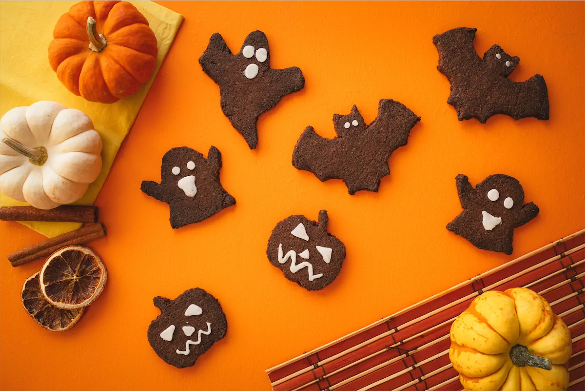 Bat, ghost, and pumpkin-shaped keto Halloween cookies are arranged on an orange table alongside red, yellow and white miniature pumpkins.