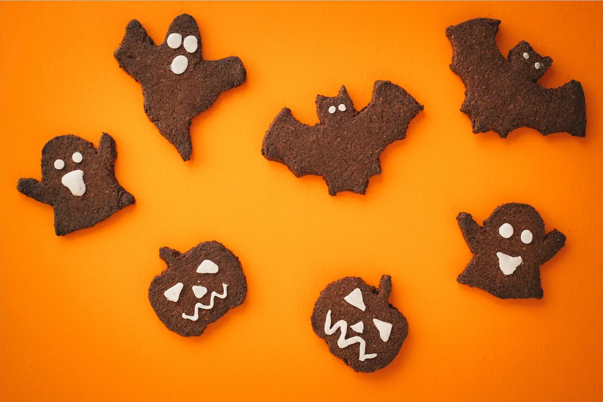 Bat, ghost, and pumpkin-shaped sugar-free keto Halloween cookies are arranged on an orange table.