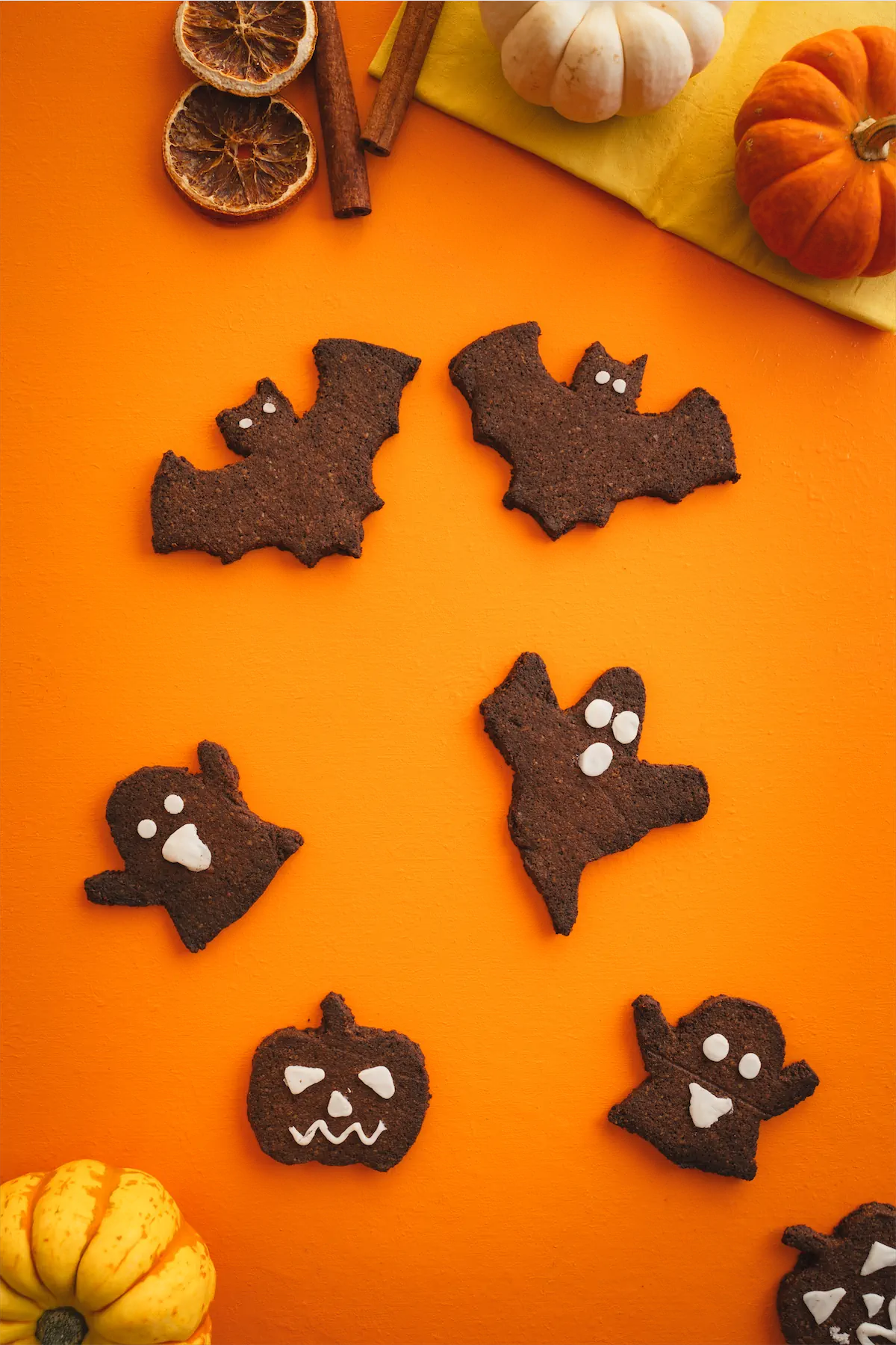 Bat, ghost, and pumpkin-shaped keto Halloween cookies are arranged on an orange table alongside red and yellow miniature pumpkins.