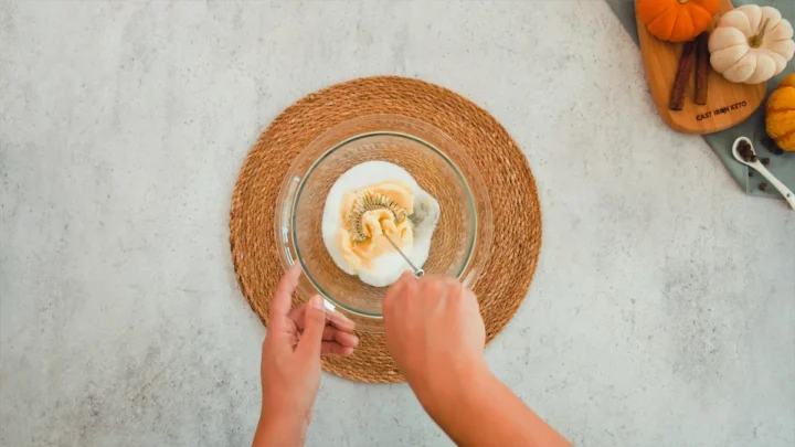 Two hands are whisking the softened butter and sweetener in a transparent glass mixing bowl.