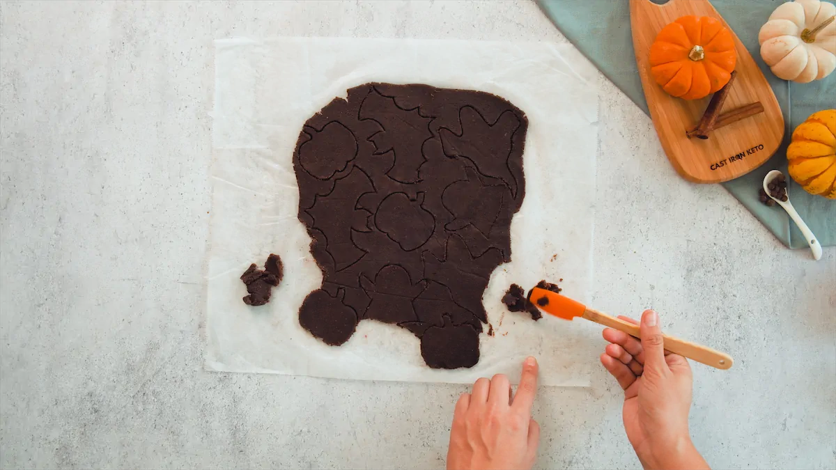 Gently lifting the carved spooky shapes with a silicone spatula from the rolled-out cookie dough.