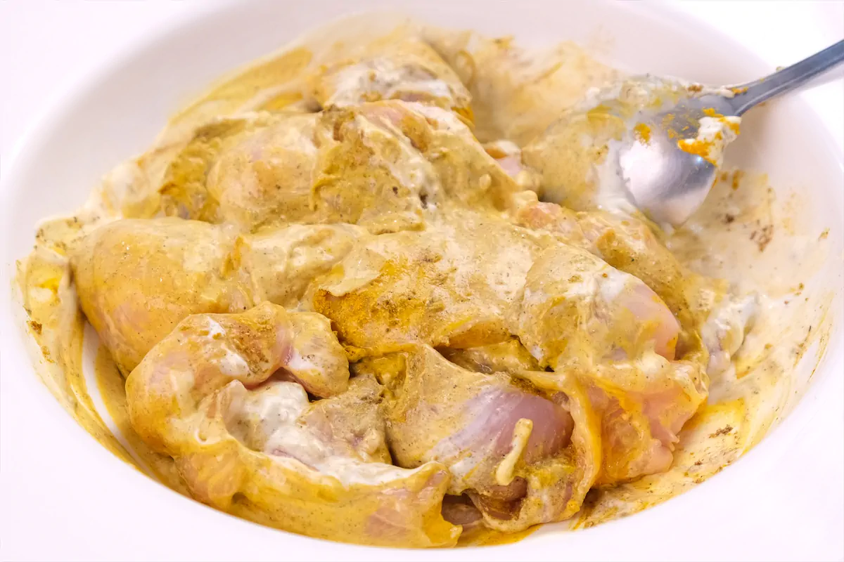 Marinating chicken pieces in heavy cream, garam masala, turmeric powder, and salt with a spoon in a bowl.