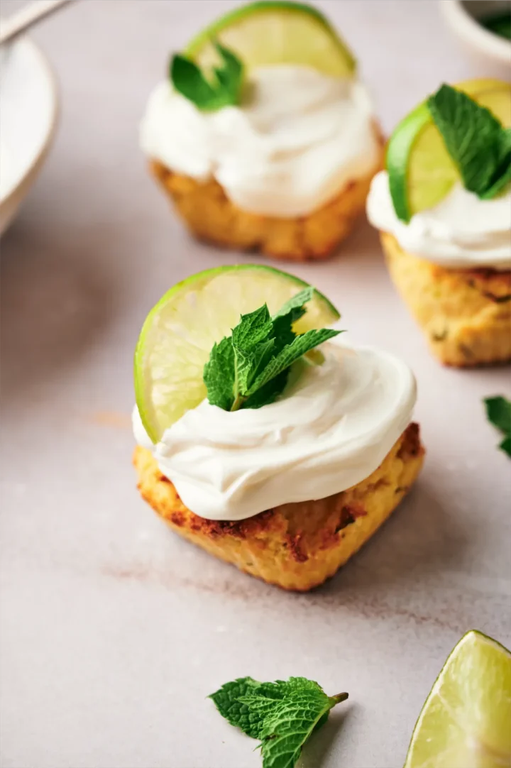 Keto mojito cupcakes, adorned with cream cheese frosting, a lemon wedge, and fresh mint leaves arranged on a table.