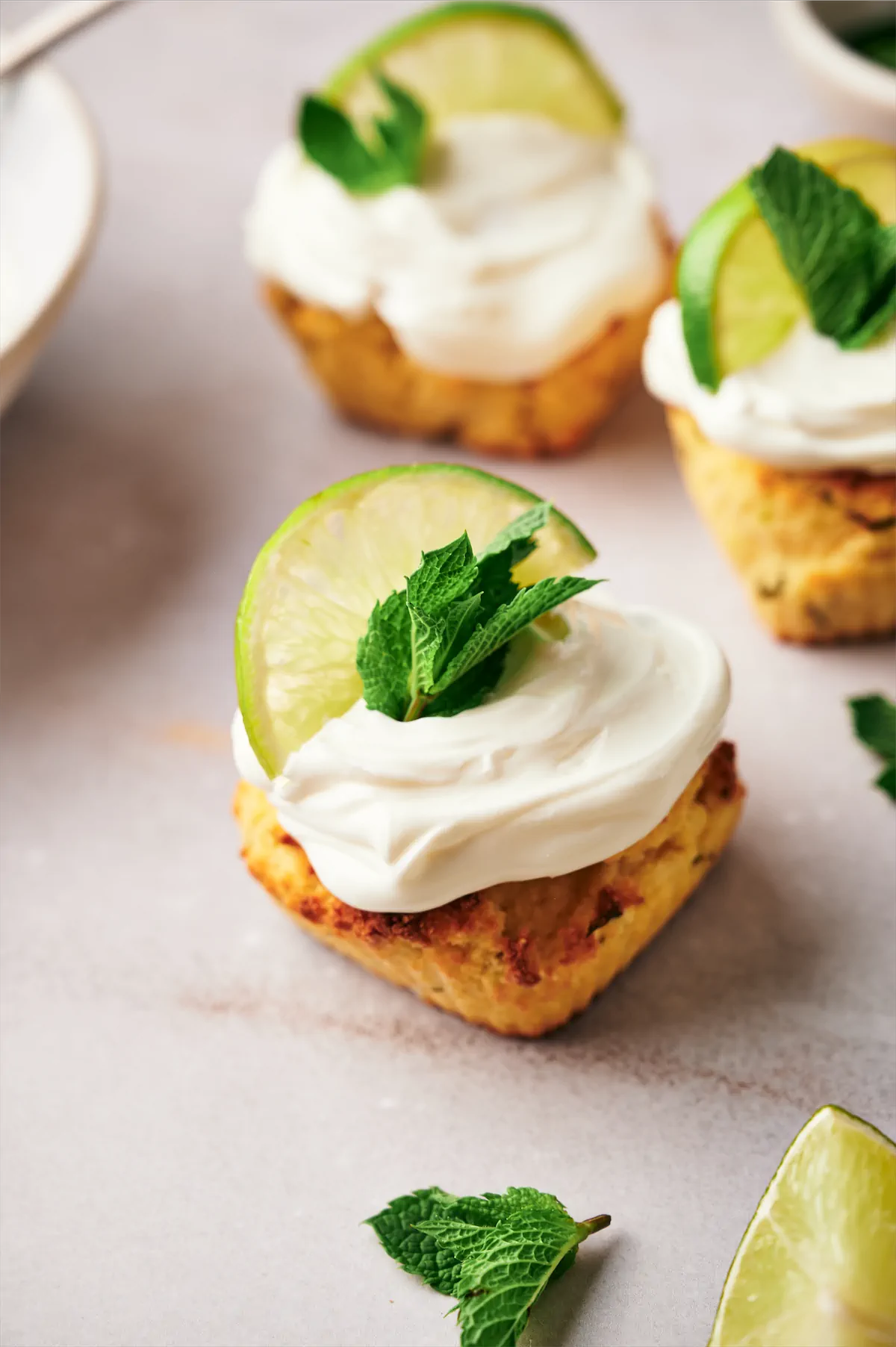 Keto mojito cupcakes, adorned with cream cheese frosting, a lemon wedge, and fresh mint leaves arranged on a table.
