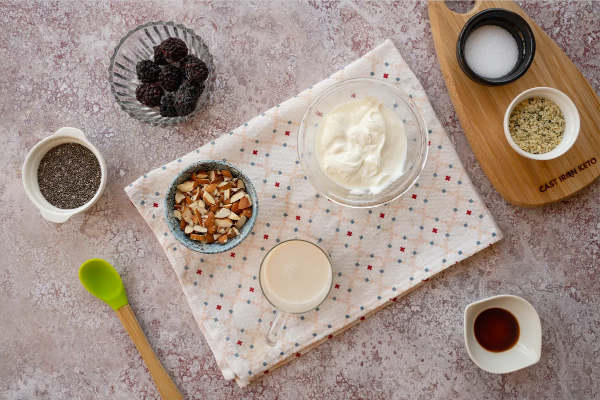 Yogurt, almond milk, chia seeds, fresh berries, almonds, and other ingredients required to make this recipe are gathered and arranged on the table.