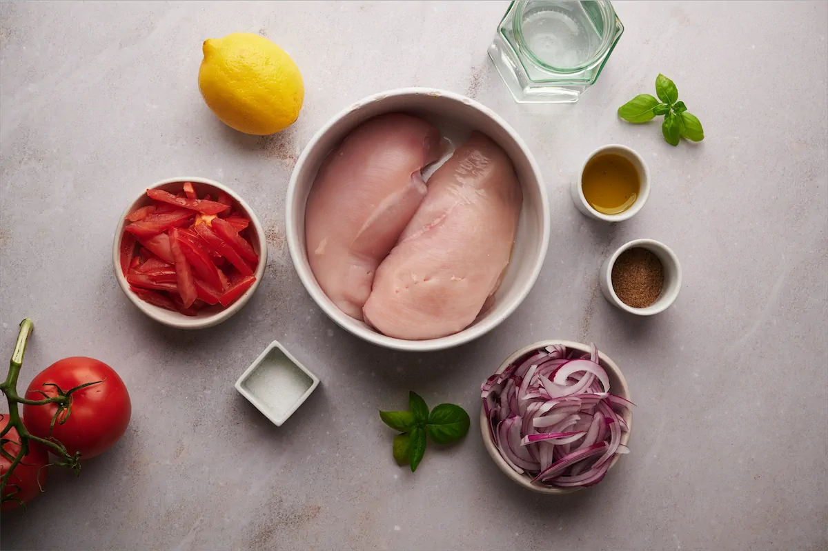 The required ingredients including olive oil, sliced red onion, thinly sliced tomato, Cajun seasoning, boneless skinless chicken breast, water, and lemon gathered a displayed on the table.