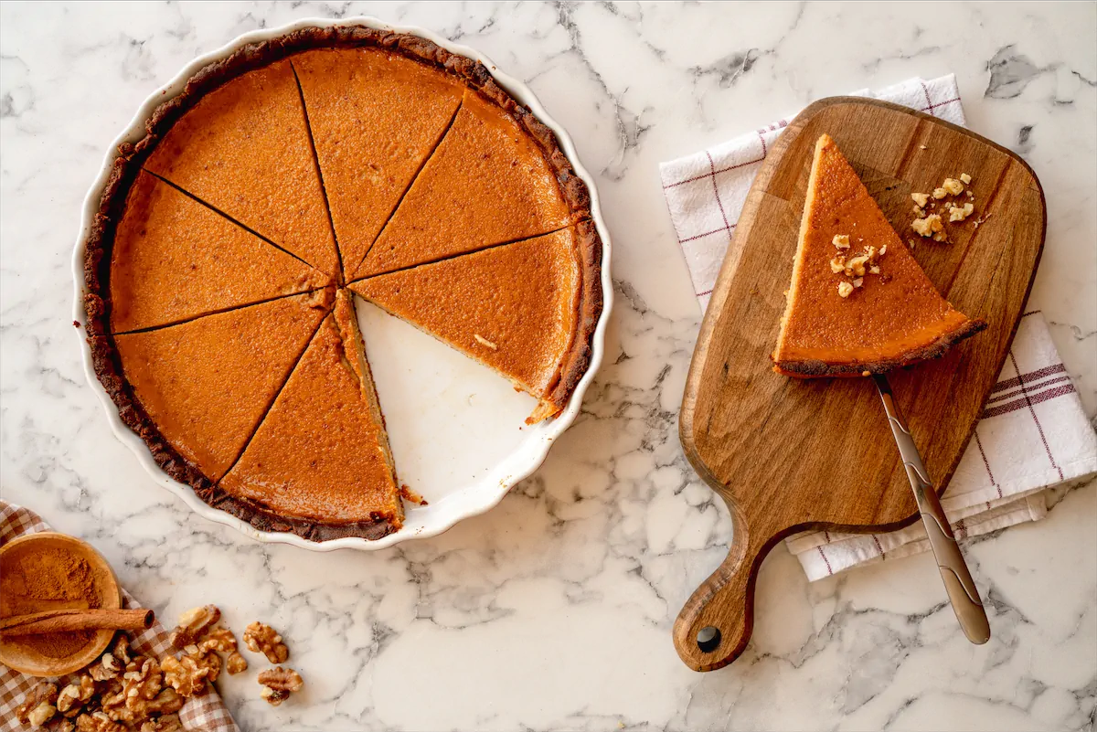 A slice of pumpkin pie topped with nuts placed on a wooden board with a server spoon alongside sliced keto pumpkin pie in a pie dish.
