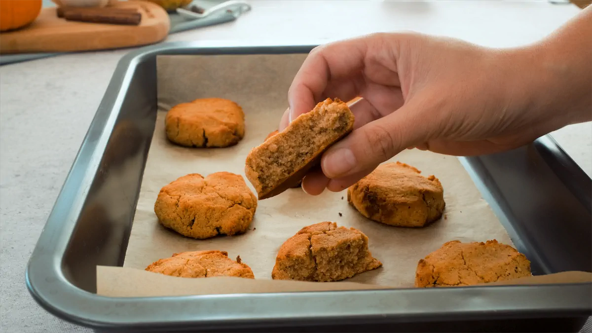 A hand holding a half a pumpkin spice cookie over a tray filled with the same cookies.