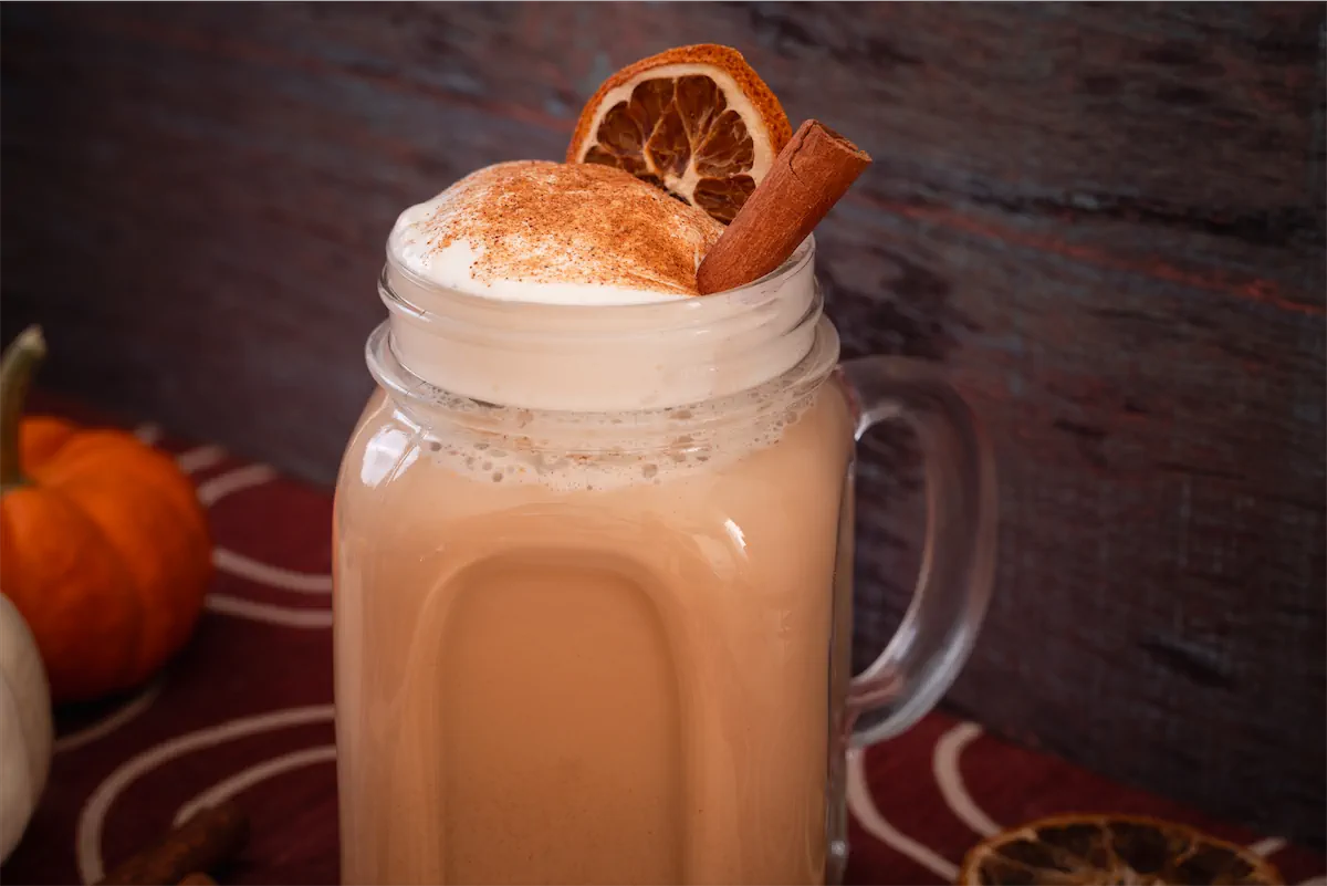 Keto pumpkin spice latte served in a mason jar finished with whipped cream, a dash of pumpkin spice, a dried orange slice, and a cinnamon stick.