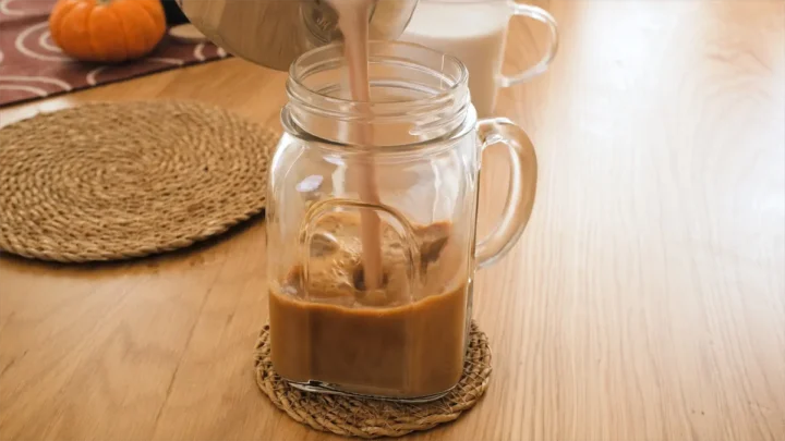 Pouring pumpkin spice almond milk mixture over the brewed coffee into a mason jar.