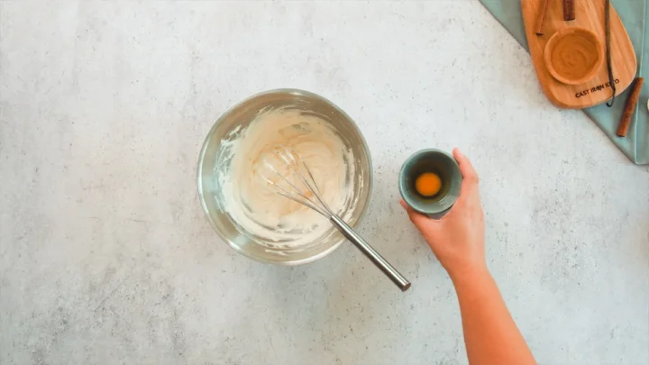 A hand holds a bowl containing a cracked whole egg, ready to pour the egg into a second bowl where butter and granulated erythritol are being whisked together.