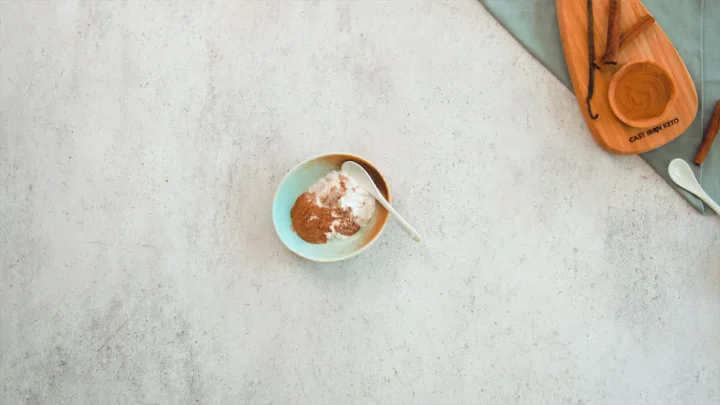 Granulated erythritol and ground cinnamon in a small bowl with a spoon ready to be mixed.