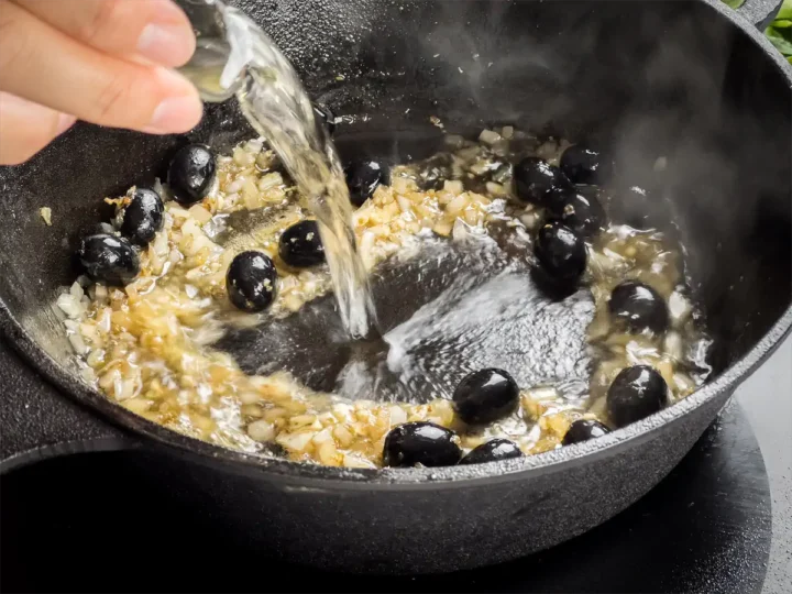Adding white wine to the cast iron skillet with aromatics and olives.