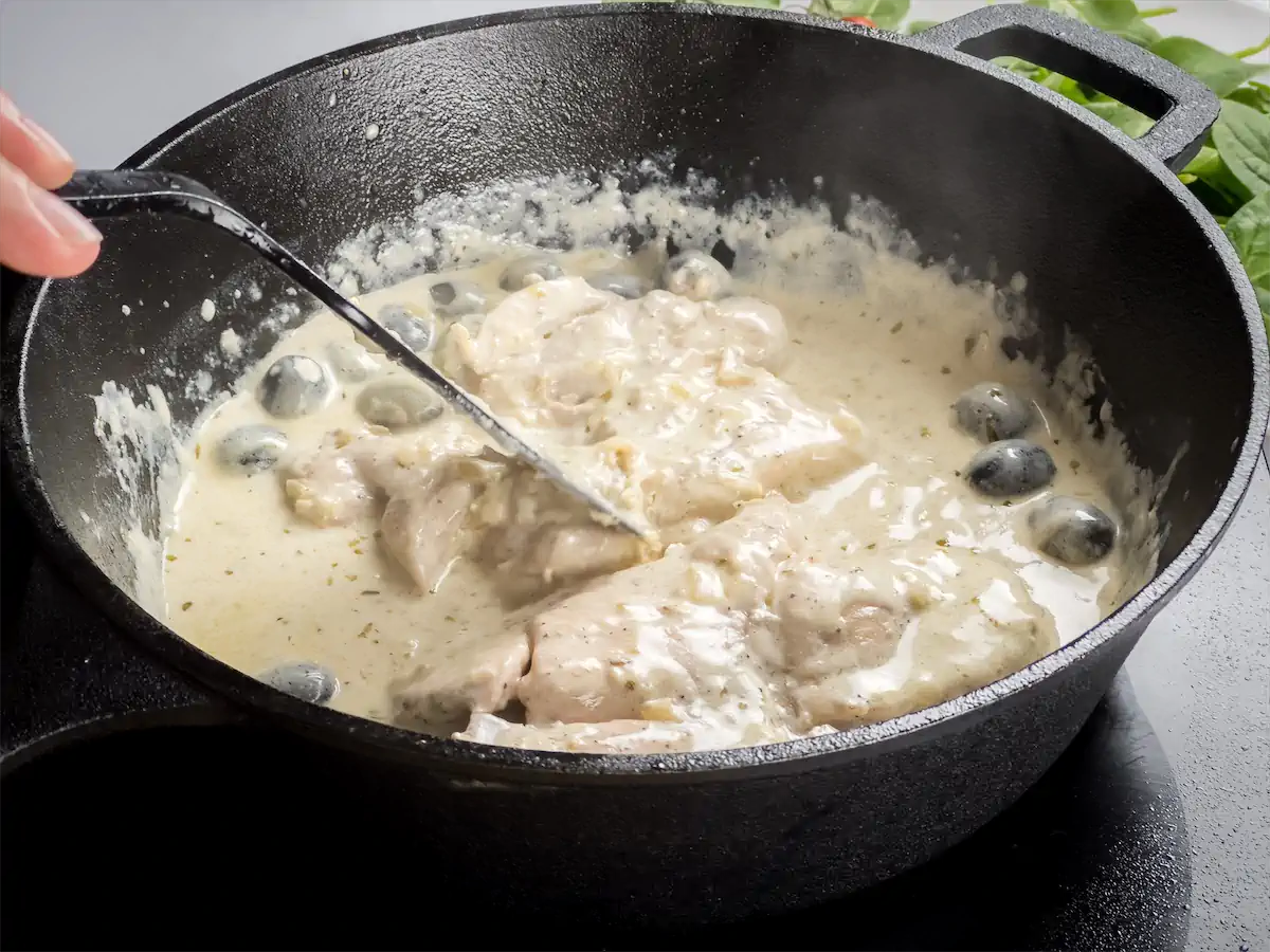 Coating the chicken pieces with creamy gravy of Tuscan chicken in a cast iron skillet with a spatula.