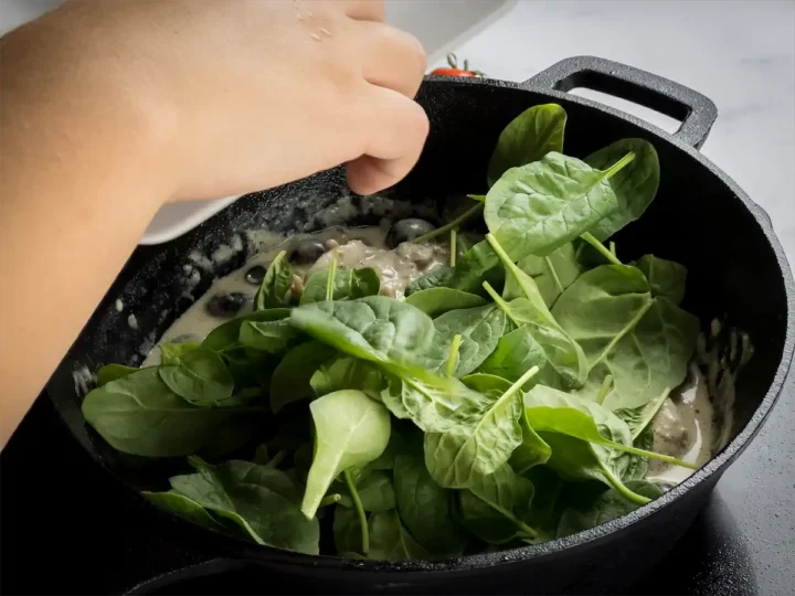 Adding the fresh spinach to the cast iron skillet with the chicken in creamy gravy.
