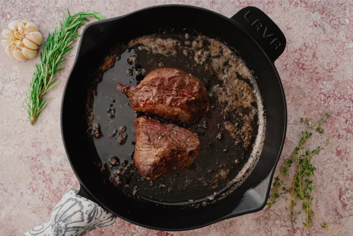 Two steaks of oven-roasted beef tenderloin with garlic butter herbs are in a cast iron skillet.