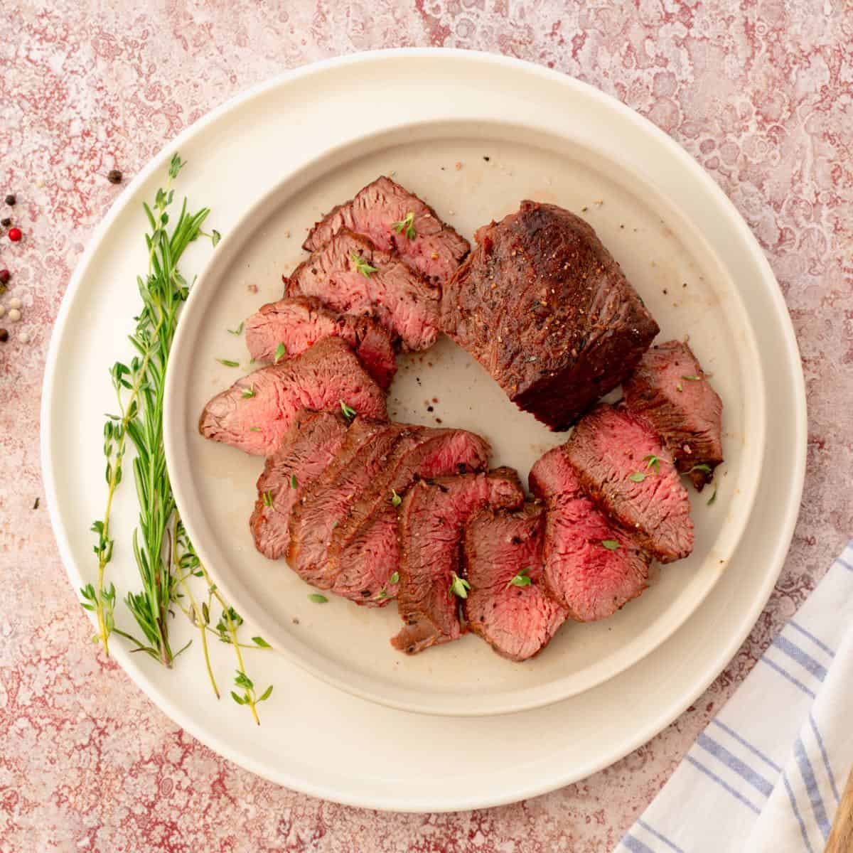 Homemade oven-roasted beef tenderloin steaks served on a white plate, a steak sliced to reveal their pink centers.