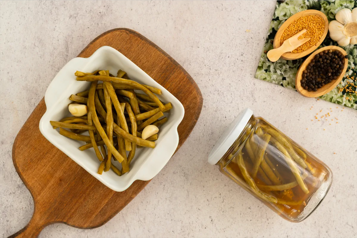 Pickled green beans are presented in a bowl, accompanied by two garlic cloves and a jar of the same pickles.