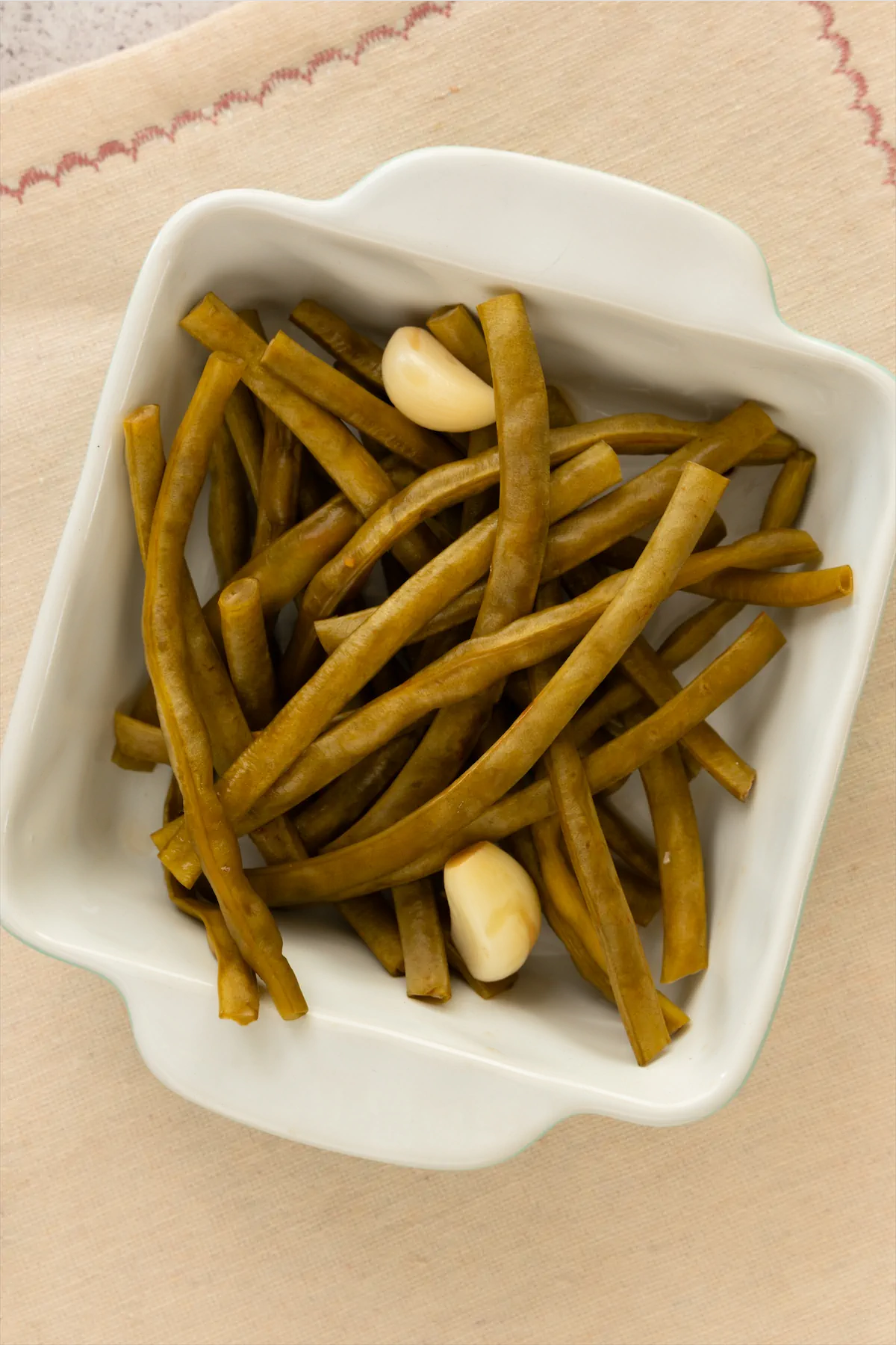 A square white bowl containing homemade pickled green beans, along with a couple of garlic cloves.