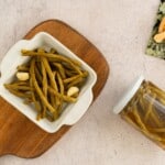 Pickled green beans served in a bowl with two garlic cloves alongside a jar of same pickles.