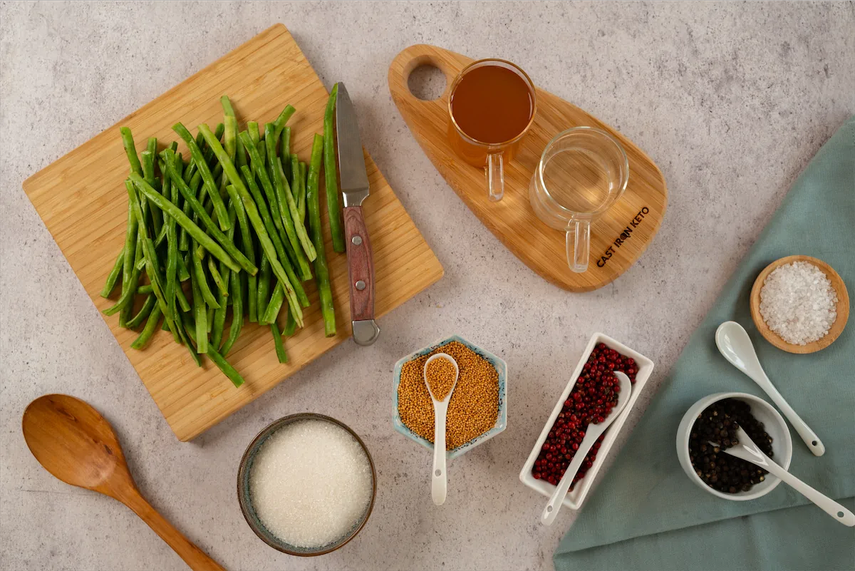 Ingredients including fresh green beans, water, apple cider vinegar, salt, whole black peppercorns, mustard seeds, red peppercorns and keto-friendly sweetener arranged and displayed on the table.