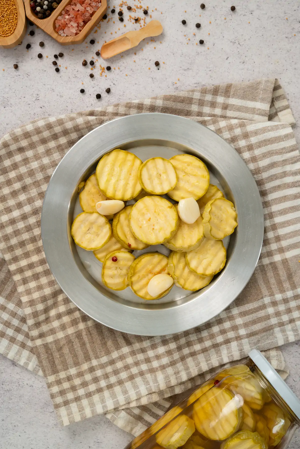 Keto sliced zucchini pickles and a couple of garlic cloves displayed on a plate alongside a jar of same pickles.