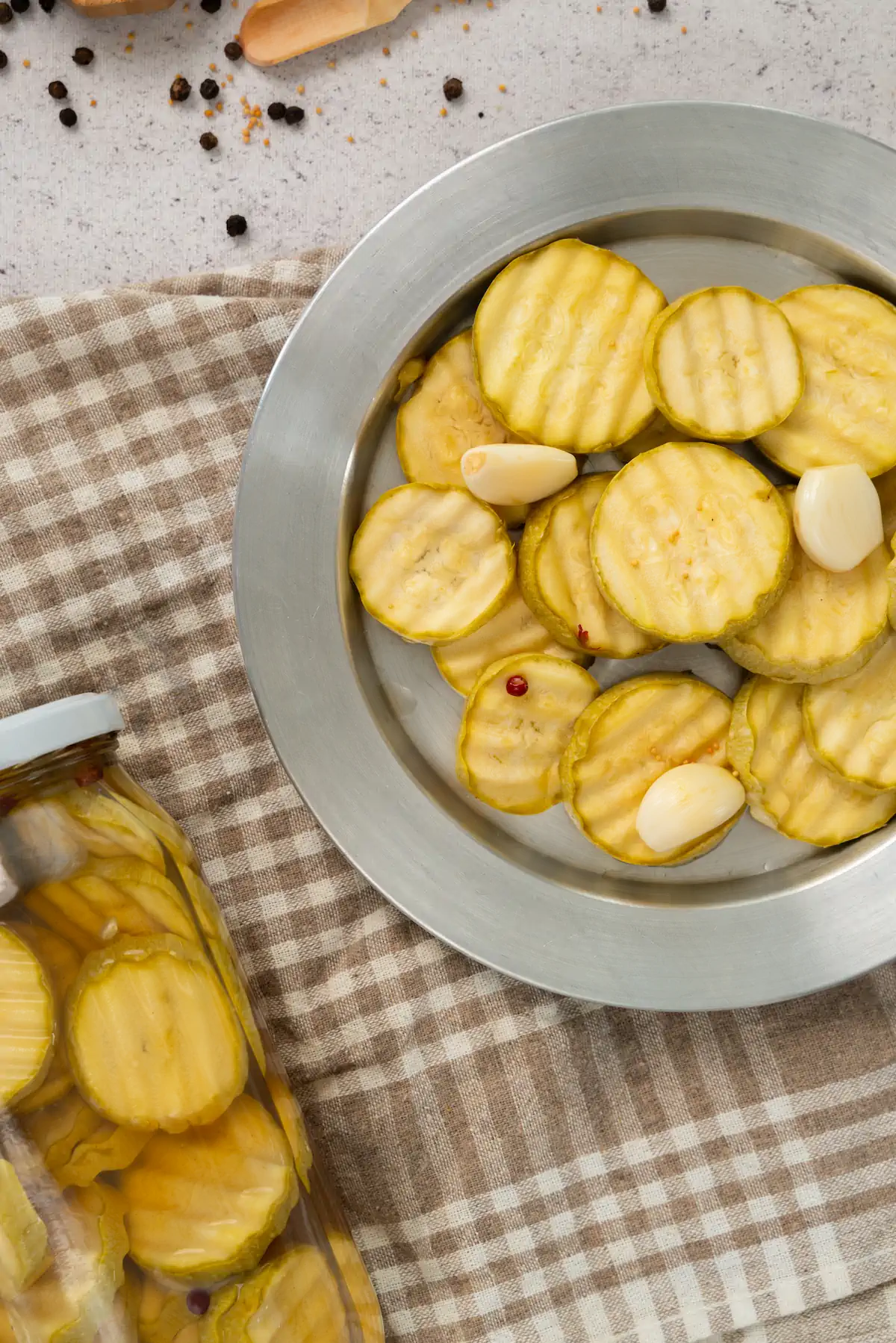 A focused shot of sliced zucchini pickles with few garlic cloves on a plate alongside a jar of same pickles.