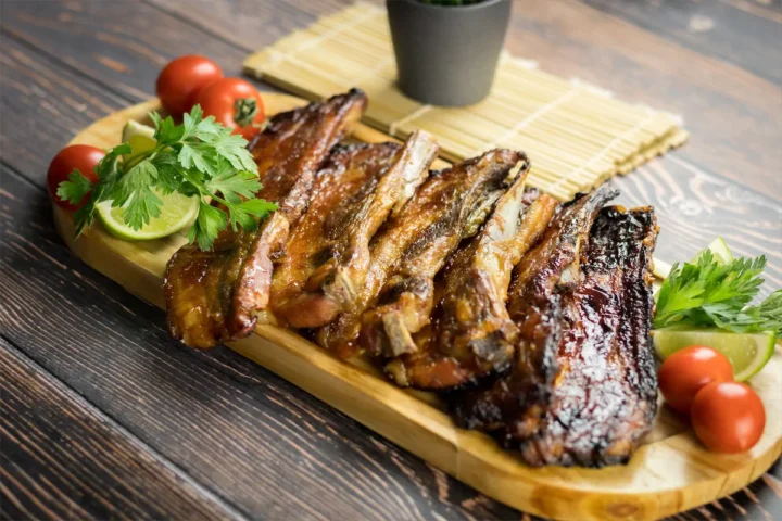 A wooden platter showcasing homemade smoked pork ribs, lemon wedge, cherry tomatoes, and fresh herbs.