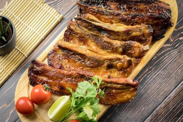 A wooden plate with homemade smoked pork ribs, garnished with lemon wedge, tomatoes, and fresh green herbs.