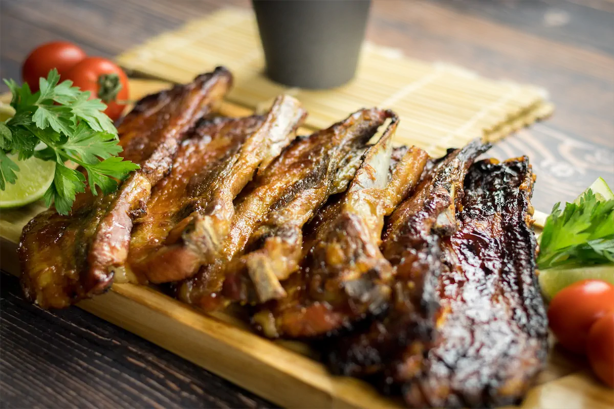 A wooden plate displaying smoked pork ribs, garnished with lemon, tomatoes, and herbs.