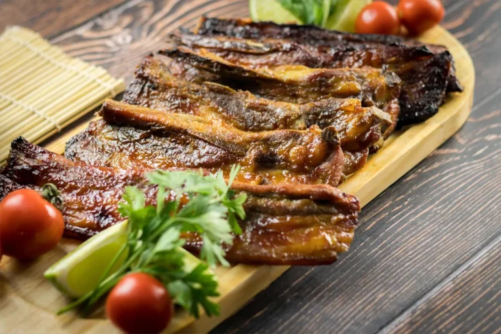 Low-carb smoked pork ribs served on wooden dish with lemon wedges, cherry tomatoes, and fresh green herbs.