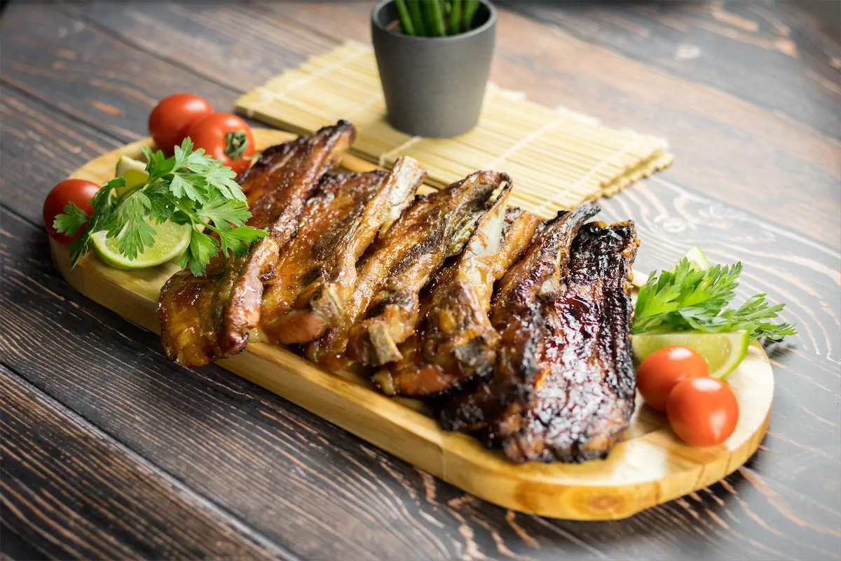 A wooden platter featuring homemade smoked pork ribs, garnished with lemon, cherry tomatoes, and herbs.