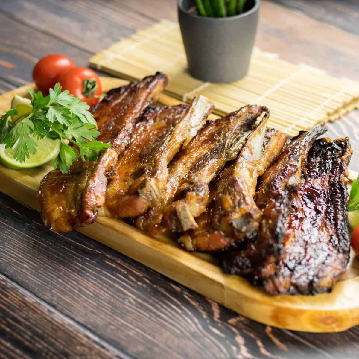 Keto smoked pork ribs presented on a wooden platter garnished with lemon wedges, tomatoes and fresh green herbs.