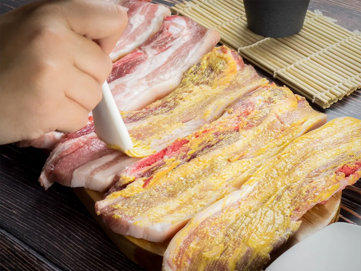 A hand brushing the pork ribs with a thin layer of yellow mustard.