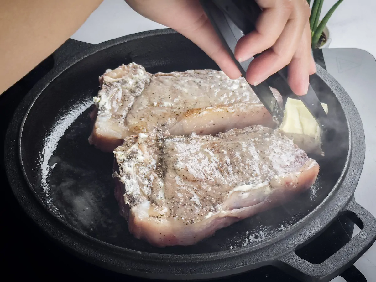 Melting a chunk of butter with tongs on a cast iron skillet with ribeye steak.