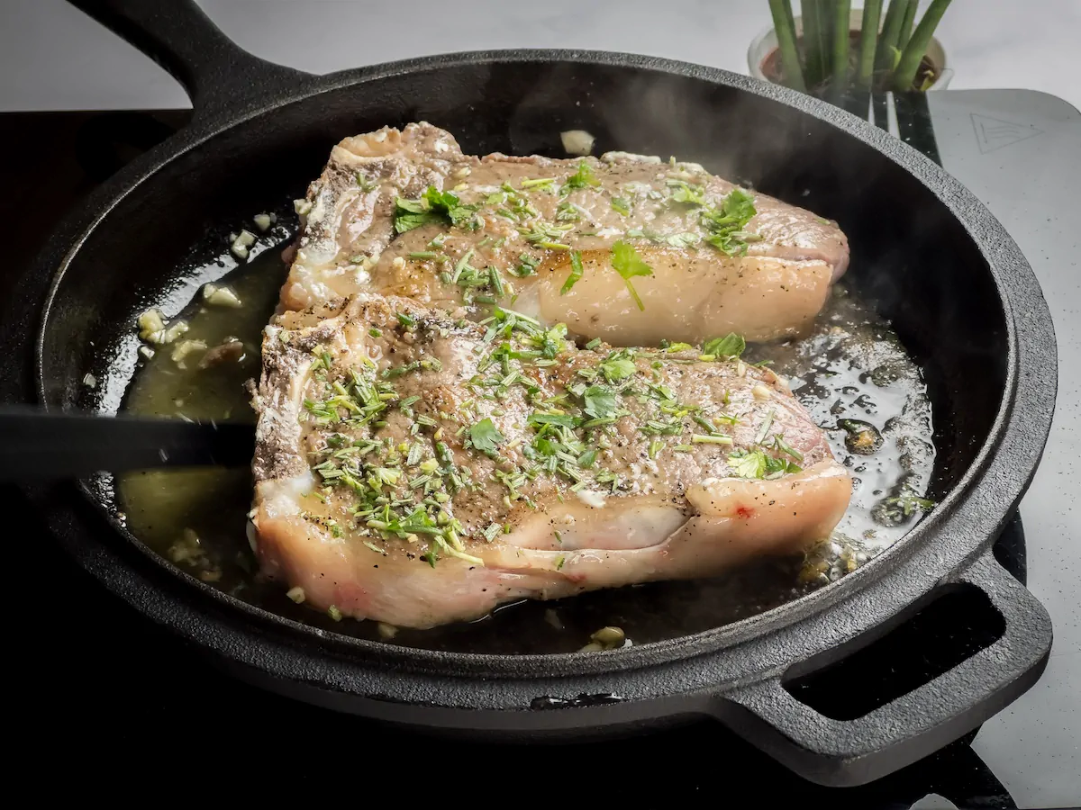 Ribeye steak with chopped fresh herbs cooking in a cast iron skillet.