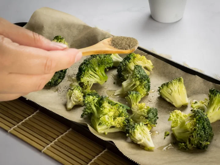 Sprinkling pepper over the broccoli florets on a baking tray lined with parchment paper.