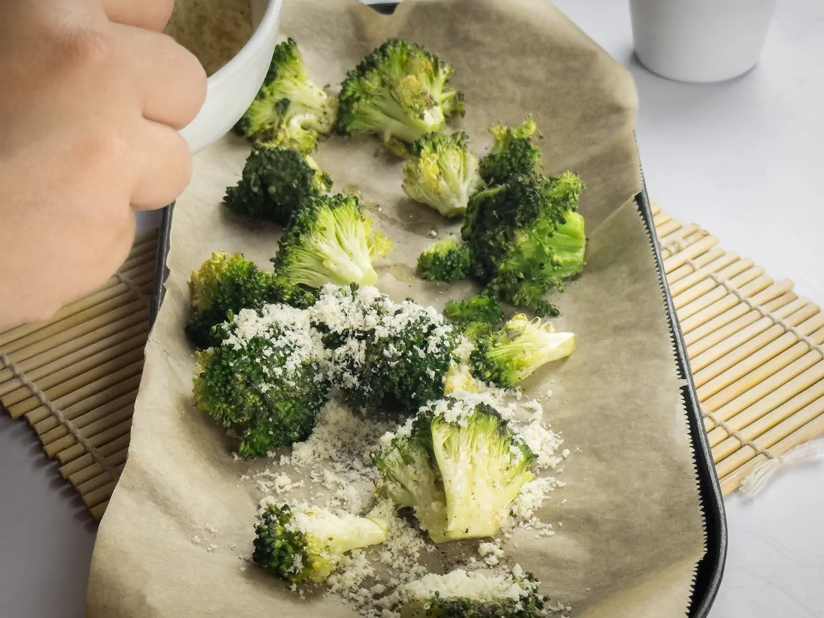 Sprinkling grated Parmesan cheese over the roasted broccoli on a baking tray.