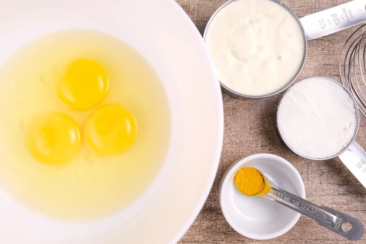 Ingredients required to make crepe batter including large eggs, heavy cream, coconut flour, and turmeric measured and gathered on the table.