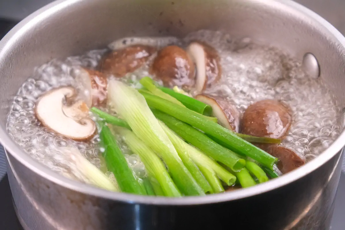 Simmering beef stock, Dashi stock granules, shiitake mushrooms, spring onions, and fresh ginger slices in a stockpot.