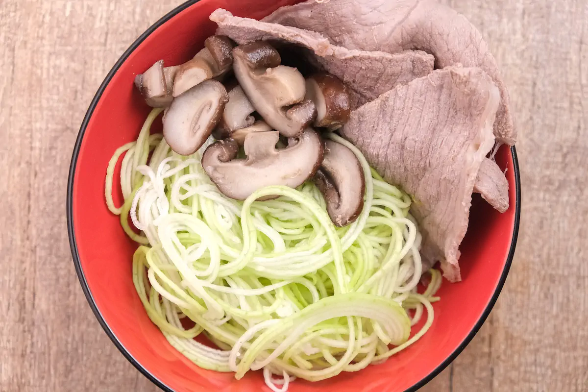 Zucchini noodles, mushrooms, and beef slices arranged in a bowl.
