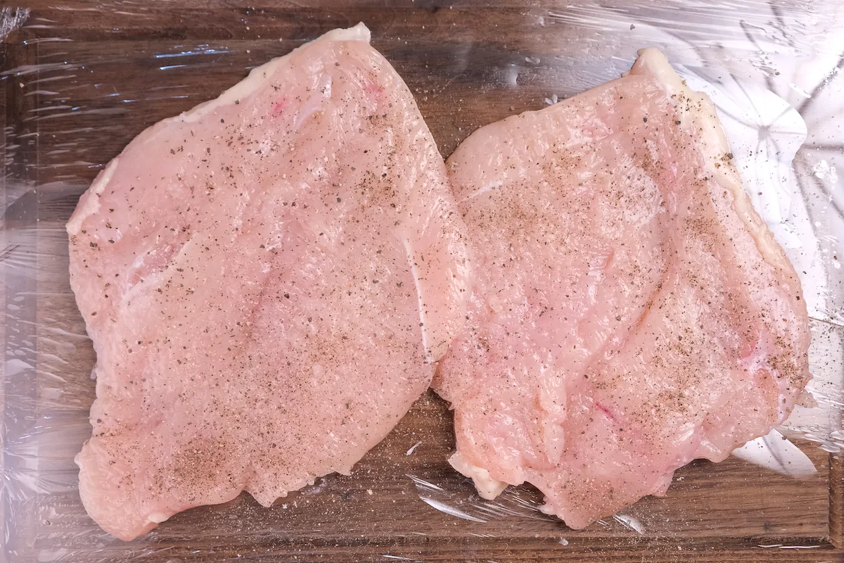 Flattened check breast seasoned with salt and pepper on a plastic wrap.