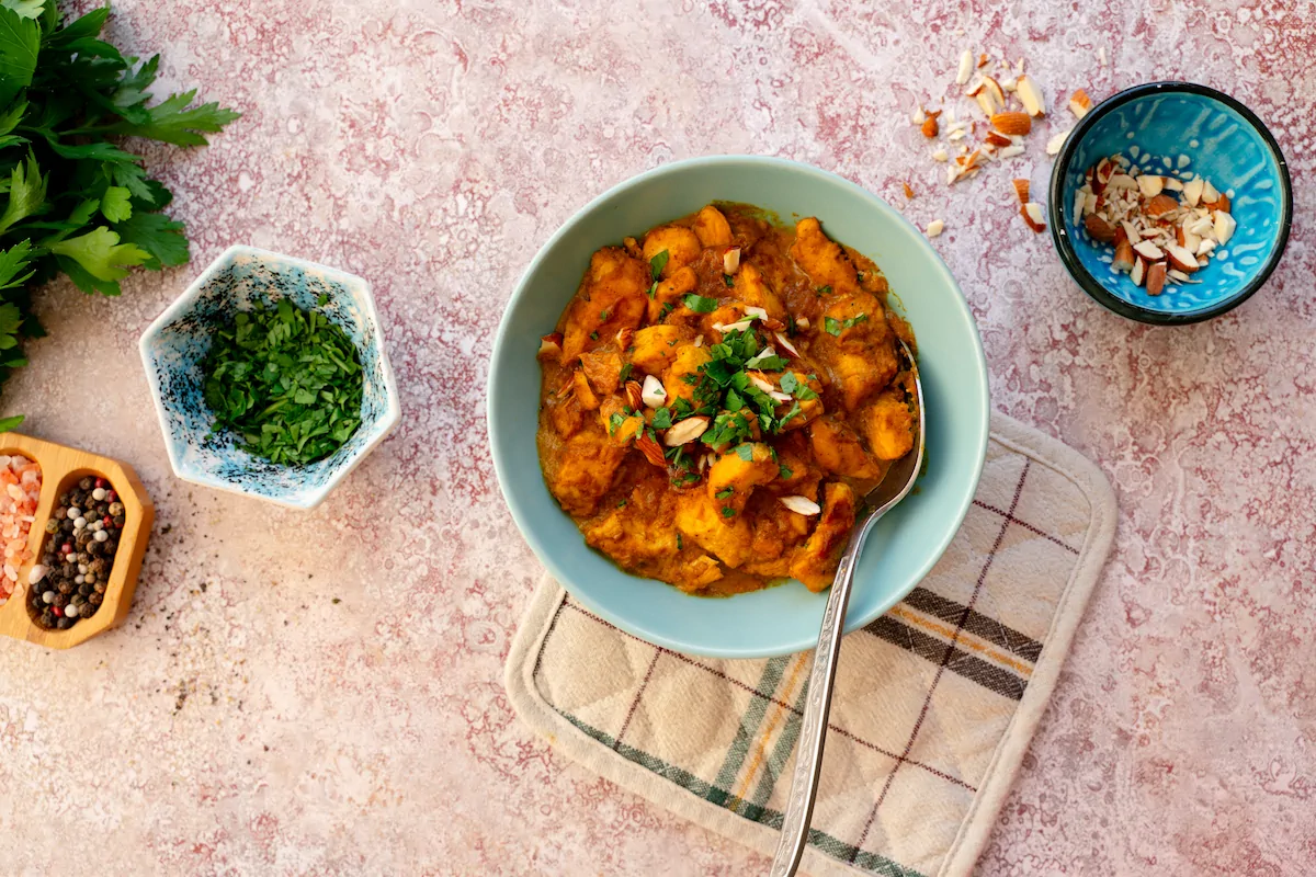 Homemade chicken korma served in a bowl garnished with green herbs and chopped nuts alongside the bowls of same garnishes.