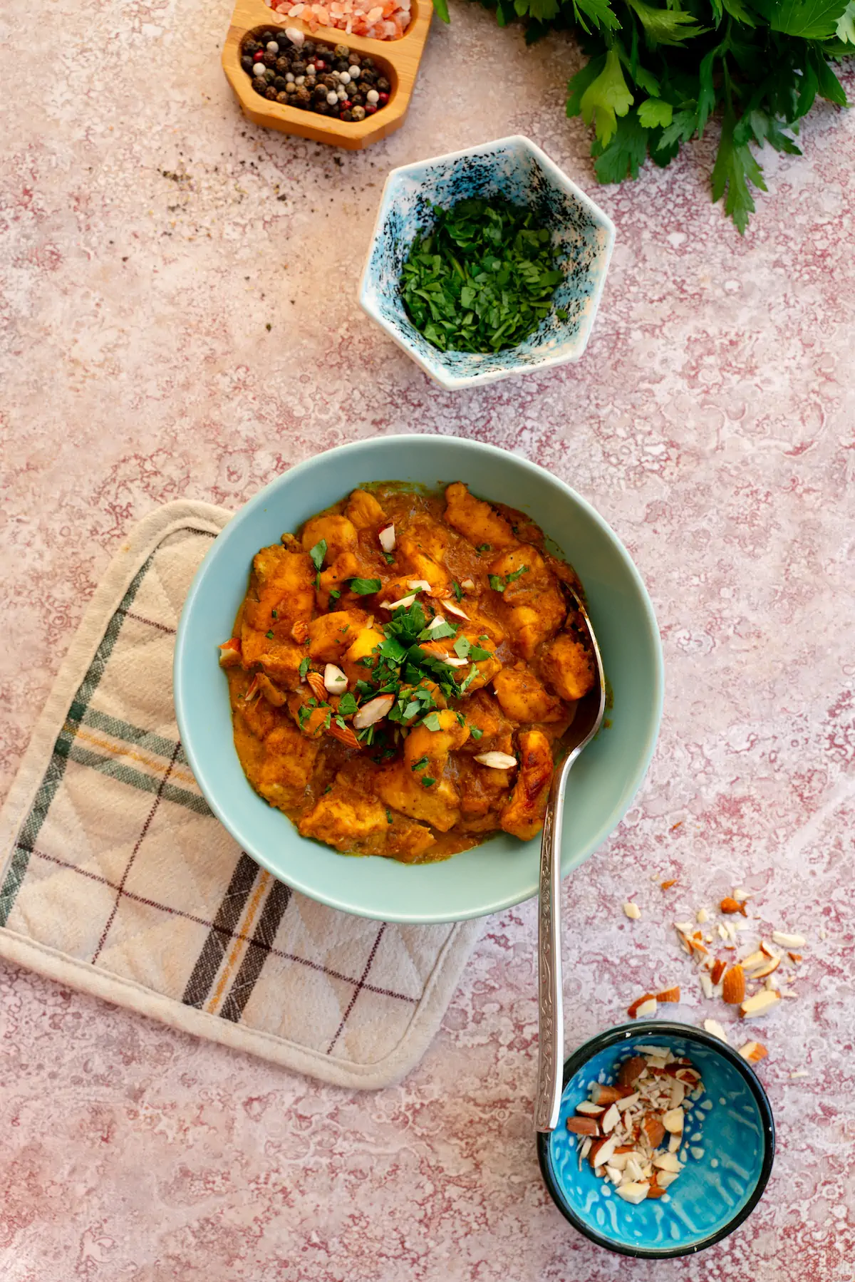 Homemade chicken korma in a bowl and served with a spoon alongside bowls of chopped fresh herbs and chopped almonds.