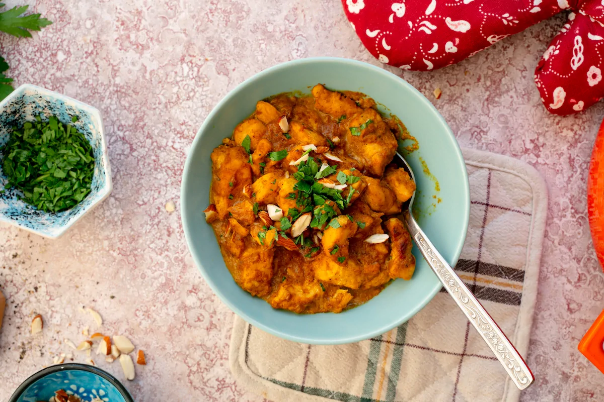 A bowl of chicken korma adorned with almonds and fresh herbs, accompanied by a spoon.