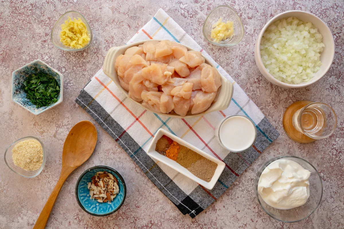 All the ingredients required to make chicken korma including skinless, boneless chicken thighs, finely chopped onion, minced garlic and ginger, full-fat plain yogurt, canned coconut milk, almond flour, spices, fresh chopped cilantro and sliced almonds gathered and arranged on the table.