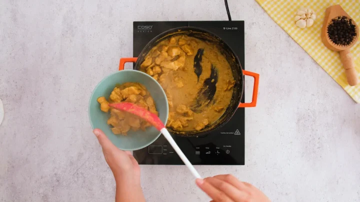 Transferring the cooked chicken korma from the cast iron skillet into a bowl with a silicone spatula.